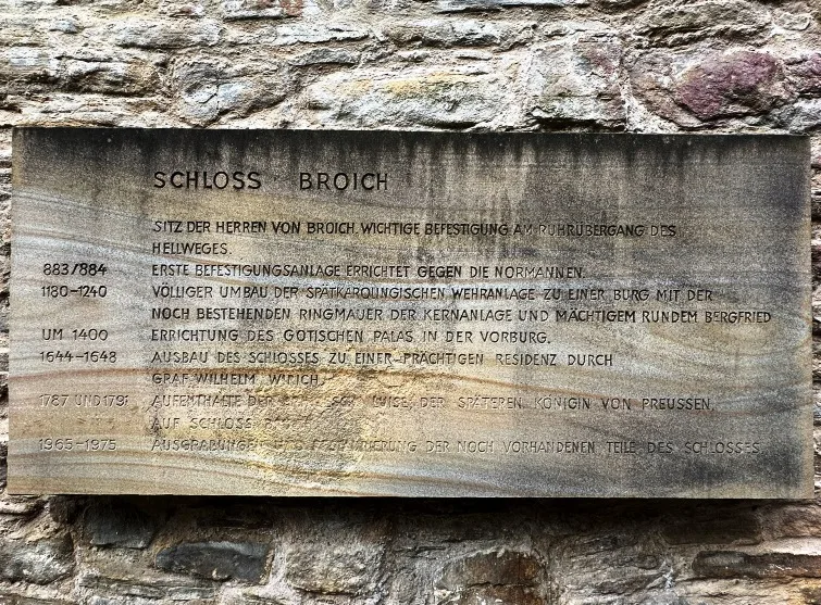 Steintafel mit historischen Informationen zu Schloss Broich in Mülheim, befestigt an der Mauer im Innenhof.