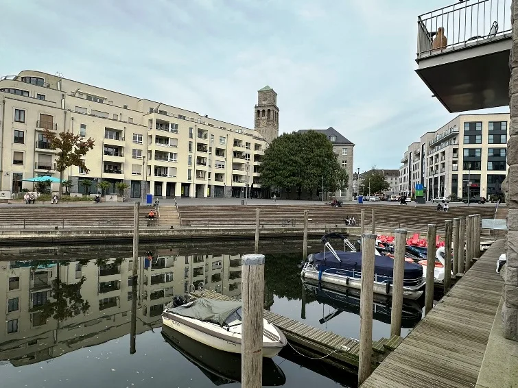 Stadthafen an der Ruhrpromenade in der Stadt Mülheim an der Ruhr, mit Blick auf moderne Gebäude entlang des Wassers und Booten im Hafen. Die Promenade ist belebt und lädt zum Spazieren und Verweilen ein.