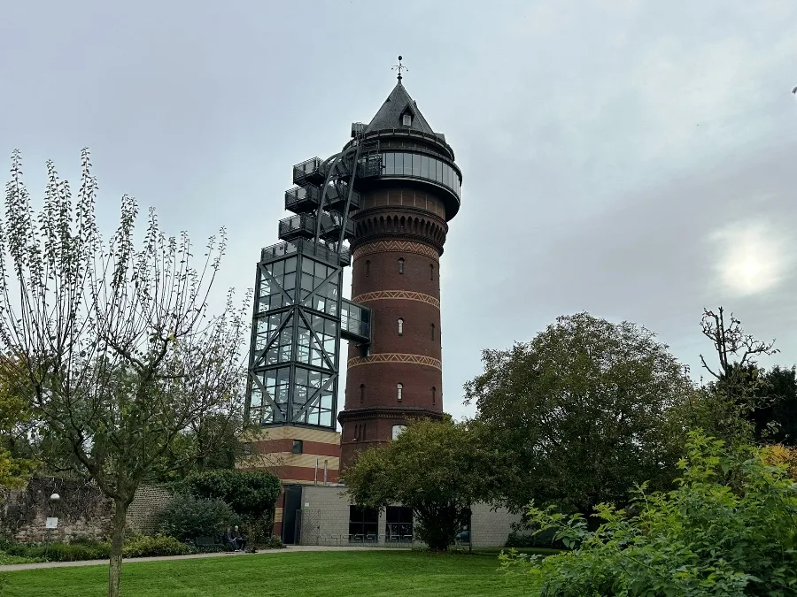 Ansicht des Aquarius Wassermuseums, ein historischer Wasserturm in Mülheim an der Ruhr, umgeben von Bäumen, unter blauem Himmel