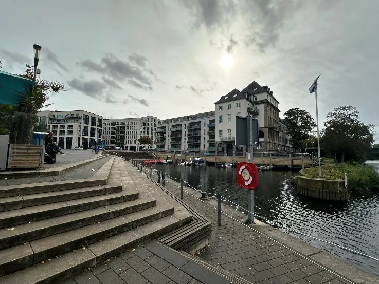 Ruhrbania in Mülheim: Blick auf den Stadthafen von Mülheim mit der Ruhrpromenade in Richtung Stadtmitte. Im Vordergrund sind moderne Gebäude und Schiffe am Wasser zu sehen, während im Hintergrund die ruhige Flusslandschaft entlang der Ruhr sowie die städtische Skyline von Mülheim sichtbar sind.