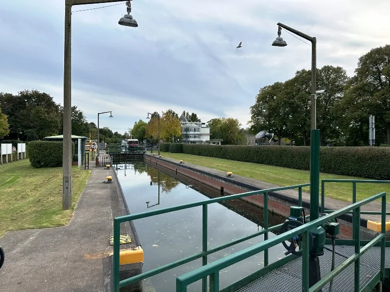 Die Schleuse auf der Schleuseninsel in Mülheim an der Ruhr, mit ihrem historischen Schleusenbecken und den markanten Schleusentoren im Vordergrund. Im Hintergrund erhebt sich der Wasserbahnhof, ein klassisches Gebäude mit seiner charakteristischen Architektur, umgeben von grüner Vegetation und Wasserflächen. Ein klarer Himmel rundet die idyllische Szenerie ab.