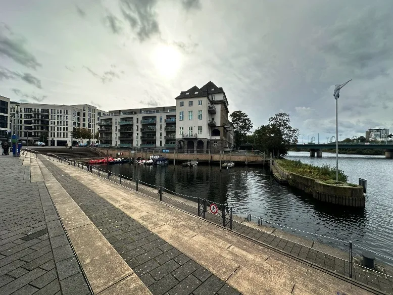 Ein stimmungsvoller Blick auf den Stadthafen in Mülheim an der Ruhr. Der Hafen zeigt eine malerische Szenerie mit ruhigem Wasser, an dessen Ufer Gebäude und Boote zu sehen sind. Im Hintergrund stehen moderne und historische Bauten, die den Charakter der Stadt widerspiegeln. Der Himmel ist wolkig, und das Licht erzeugt eine sanfte Atmosphäre.