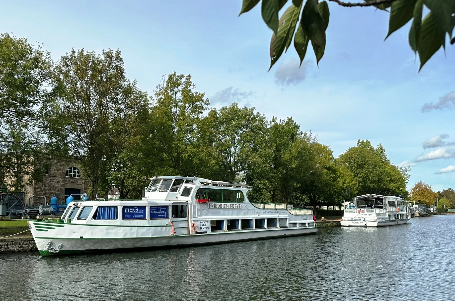 Die Weiße Flotte in Mülheim: Ein elegantes Ausflugsschiff liegt am Ufer des Wasserbahnhofs auf der Schleuseninsel, umgeben von grüner Natur.