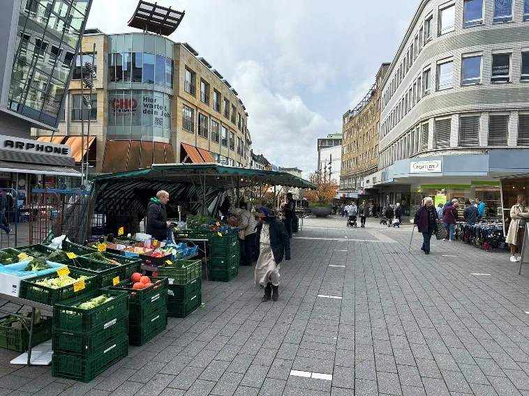 Wochenmärkte in Mülheim: Der Wochenmarkt in der Innenstadt von Mülheim an der Ruhr: Eine lebhafte Szene mit Marktständen, an denen frisches Obst, Gemüse, Blumen und andere regionale Produkte angeboten werden. Menschen schlendern zwischen den Ständen, genießen die entspannte Atmosphäre und machen Einkäufe. Im Hintergrund sind historische Gebäude und Straßenzüge der Innenstadt zu sehen.