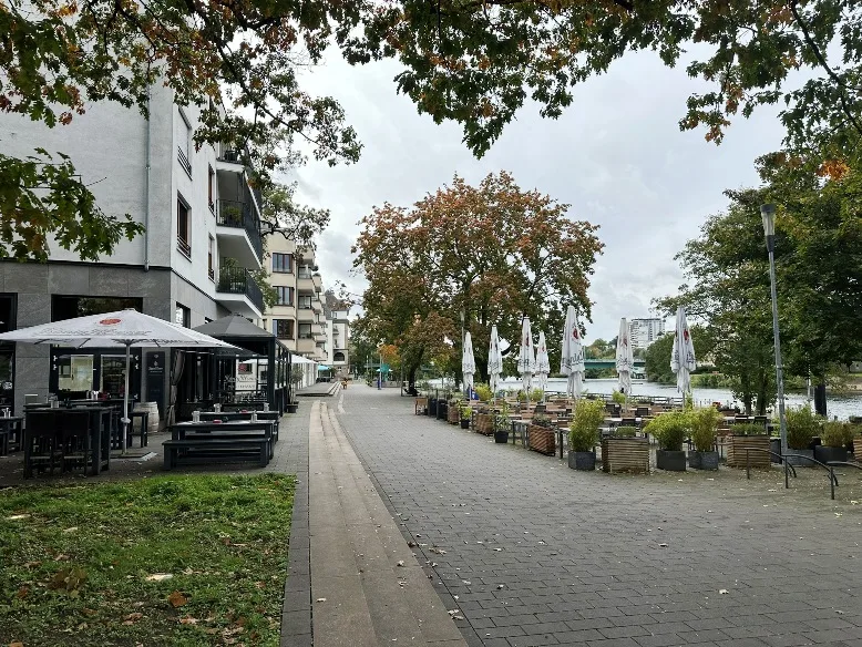 Die Ruhrpromenade in Mülheim an der Ruhr bei strahlendem Sonnenschein: Entlang des geschwungenen Uferwegs flanieren Menschen, vorbei an modernen Gebäuden und gemütlichen Cafés, die zum Verweilen einladen. Im Vordergrund spiegeln sich Boote und die grüne Uferbepflanzung im glitzernden Wasser der Ruhr, während sich im Hintergrund die urbane Skyline mit ihrem harmonischen Mix aus Natur und Architektur abzeichnet. Eine entspannte und lebendige Atmosphäre, die die Lebensqualität der Stadt am Fluss unterstreicht.